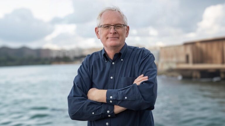 Mark Tilbury standing by the waterfront with arms crossed, wearing a navy blue button-down shirt and glasses.