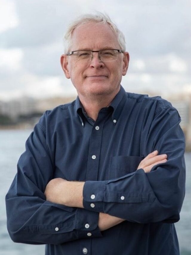 Mark Tilbury standing by the waterfront with arms crossed, wearing a navy blue button-down shirt and glasses.