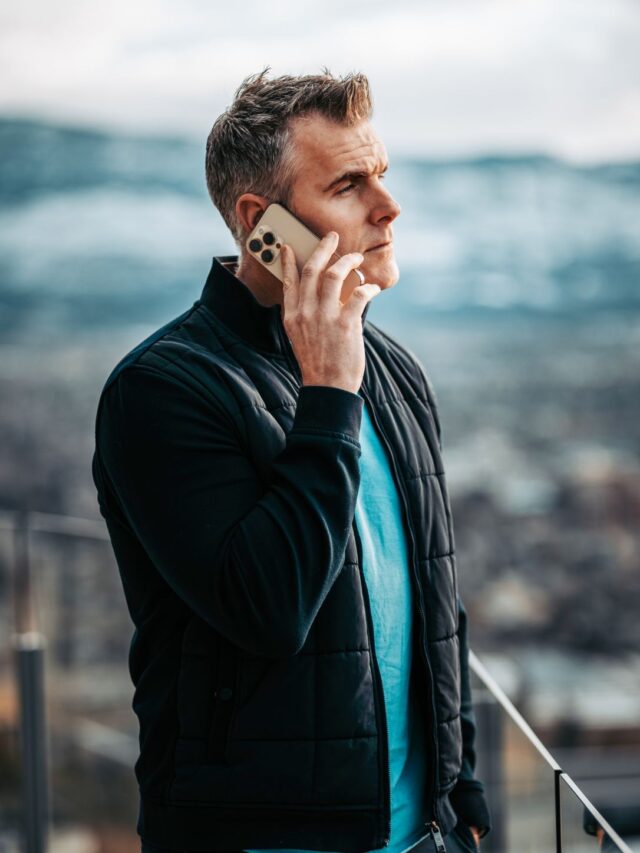 A photo of Dan Martell, a successful entrepreneur and author, talking on his cell phone. He is wearing a black jacket over a blue t-shirt and is standing in front of a city landscape.  